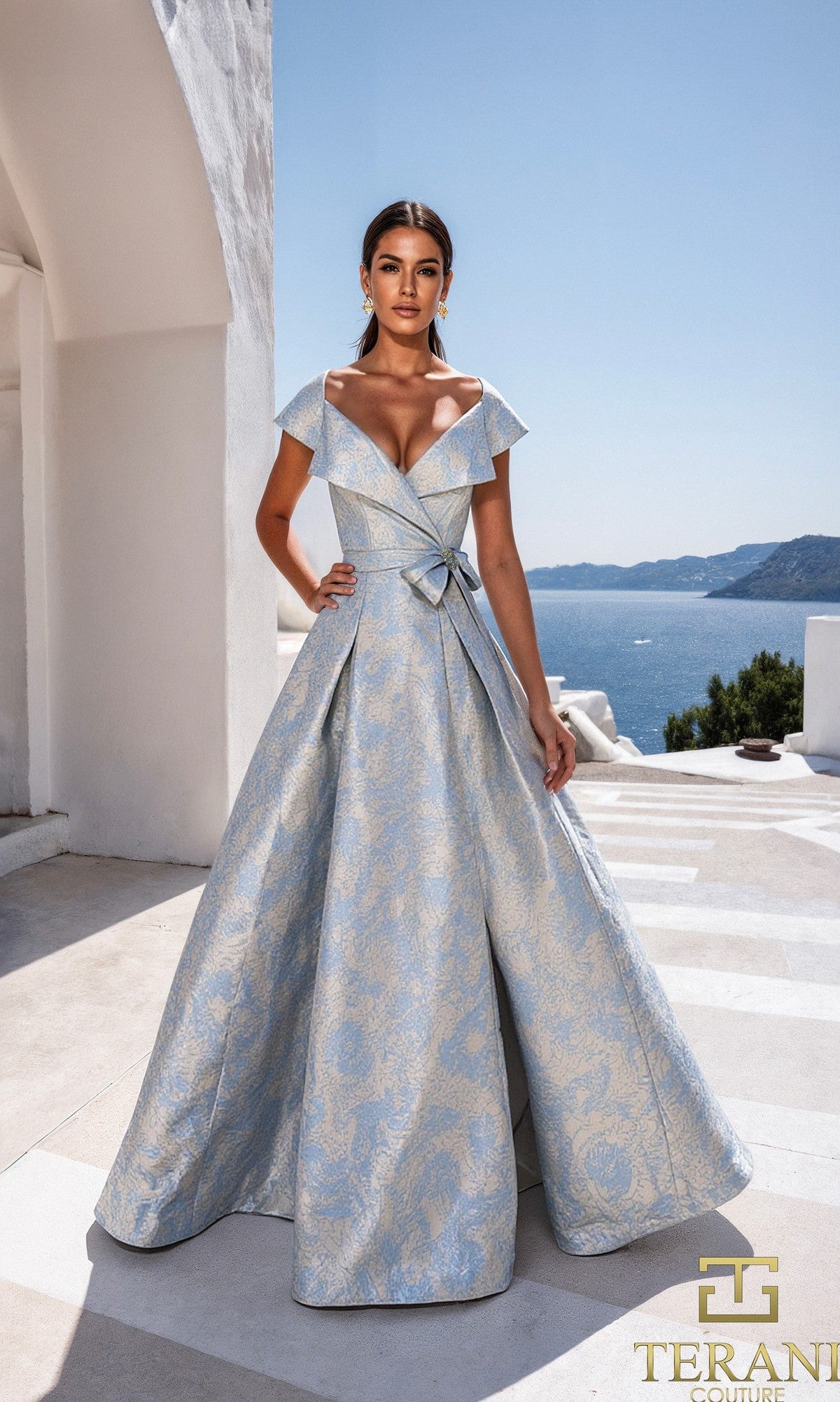 A woman in a Blue Stone Terani 261M6443 long formal dress stands outdoors on a sunlit terrace, with the sea and hills in the background.